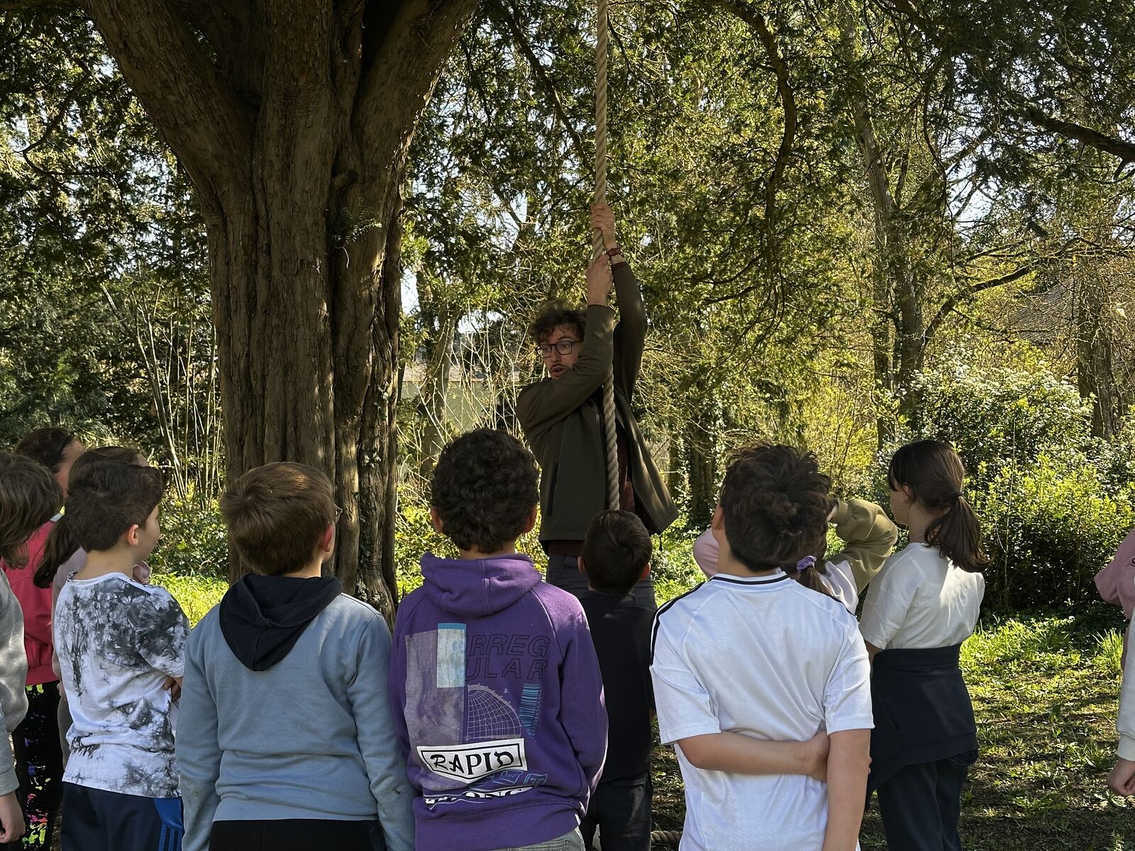 Un moniteur grimpe à une corde suspendue à un arbre pendant qu'un groupe d'enfants regarde